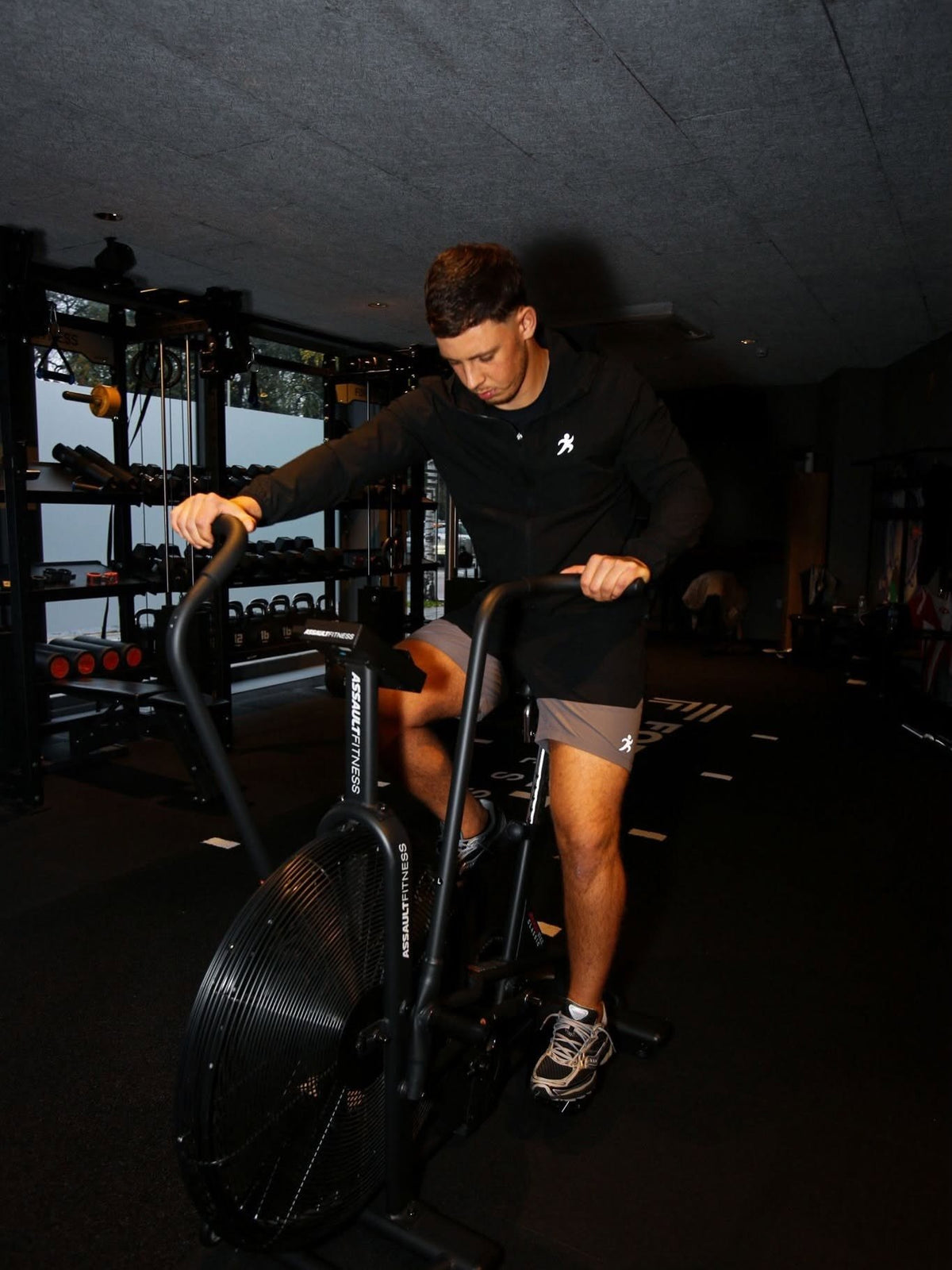 Training on an air bike in a black jacket, grey shorts and Saucony trainers
